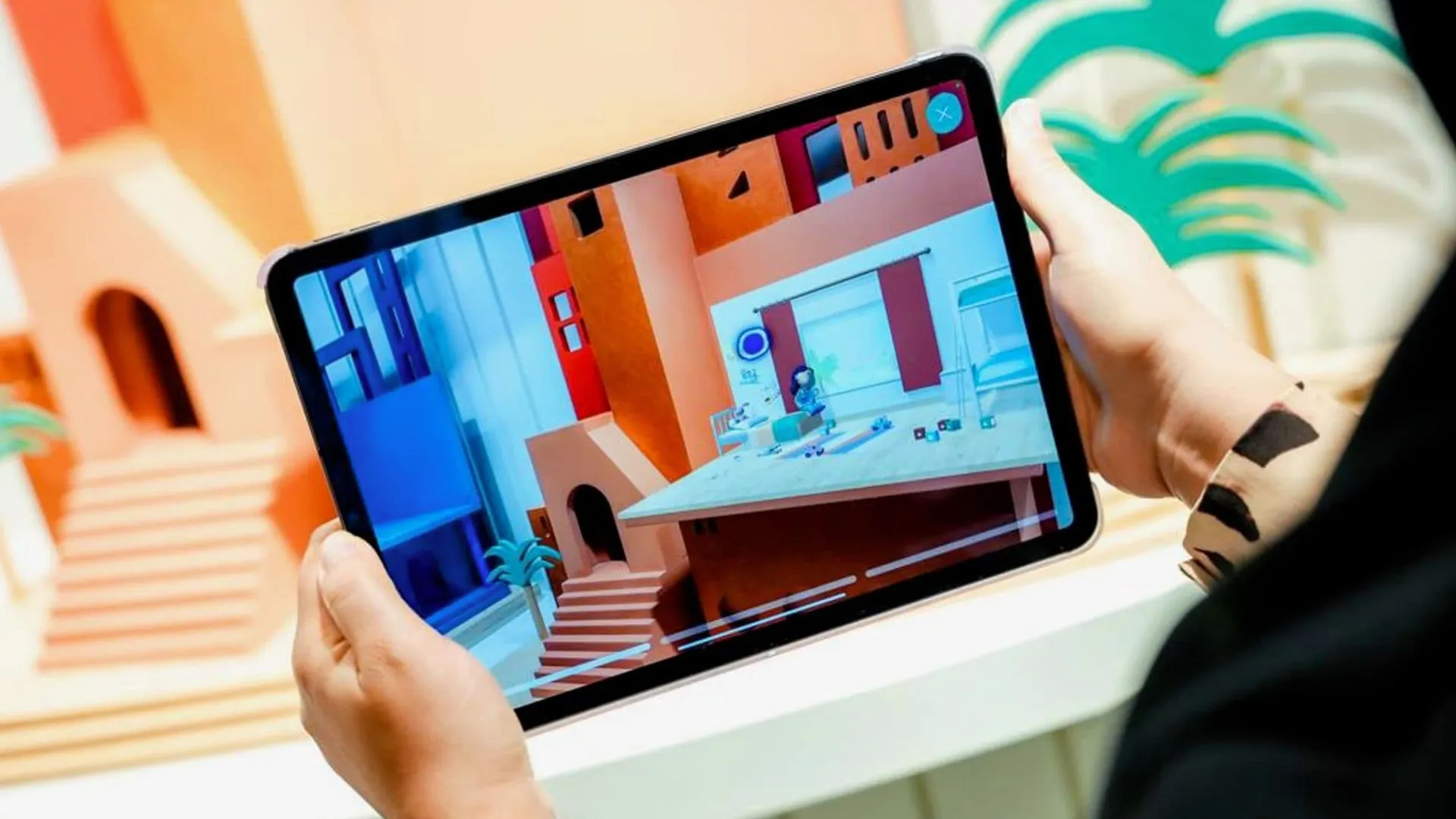 A hand holding a tablet showing a colourful child's-eye view of a town
