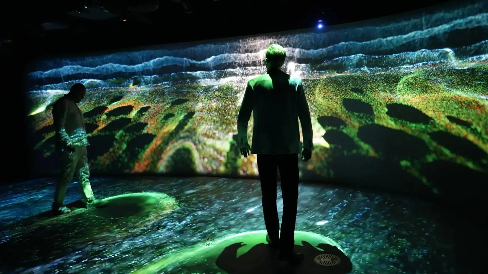 Visitors standing inside the immersive projection room, surrounded by a glowing landscape