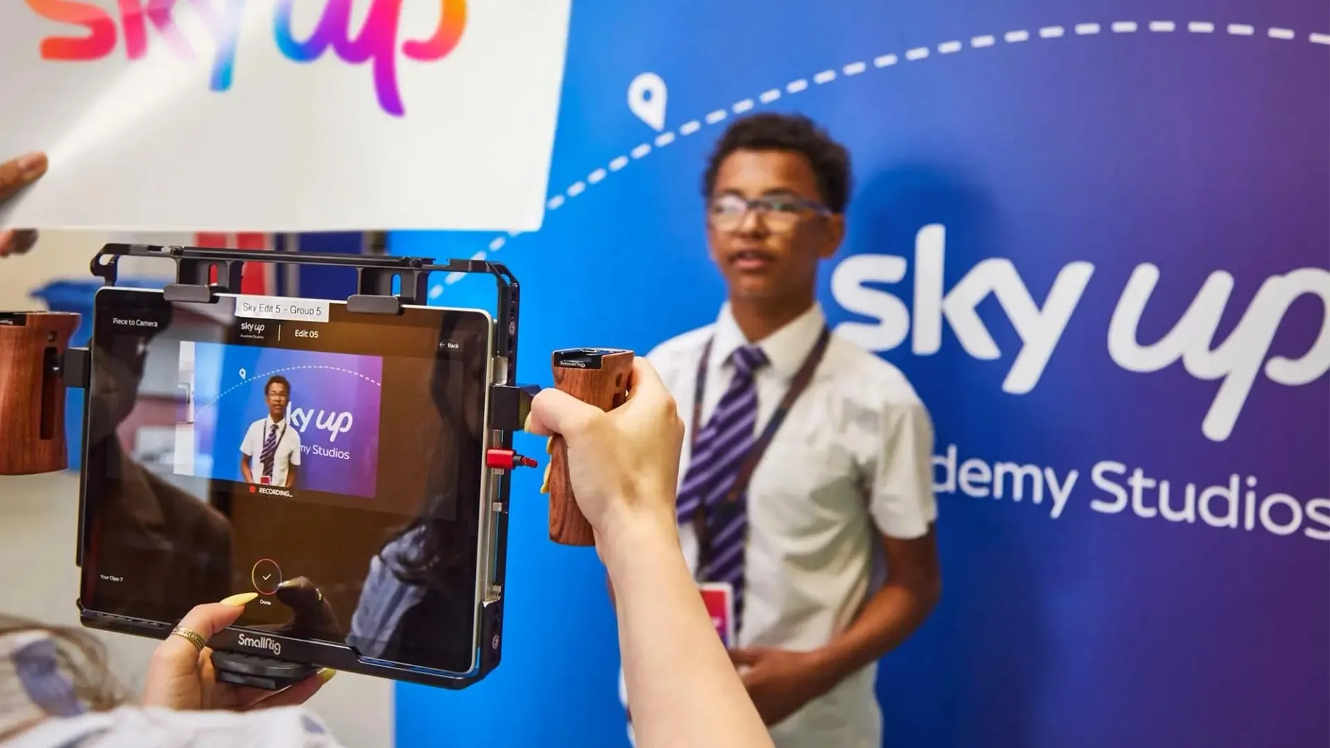 A student filming a classmate on a handheld camera rig in front of the Sky Up Academy Studios wall