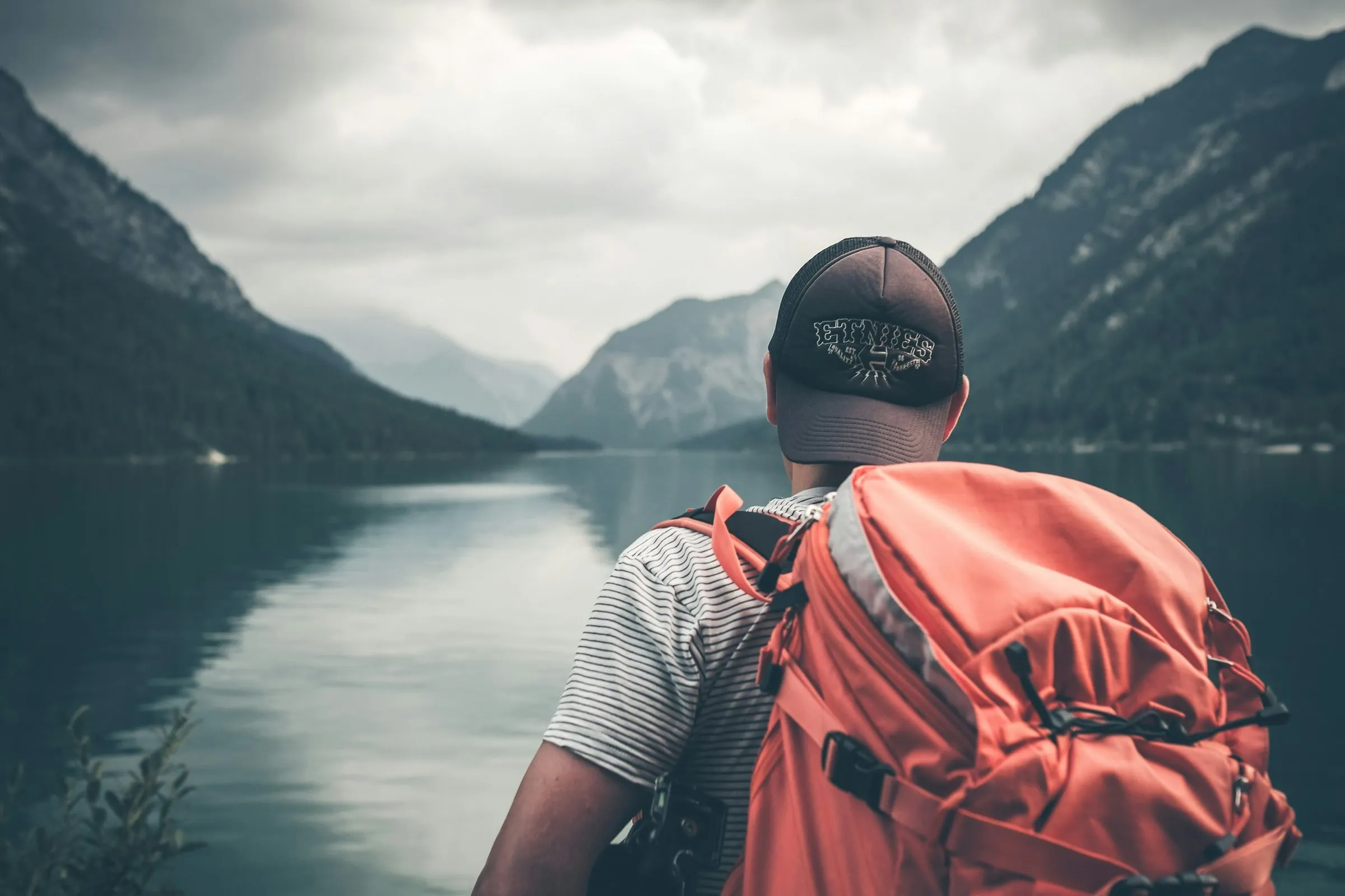 A traveller with an orange backpack looking out over a mountain lake
